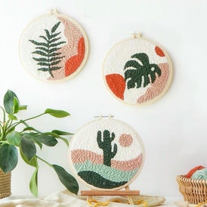 Pode incluir: Tr&ecirc;s aros de bordado com diferentes desenhos inspirados na natureza. O primeiro aro apresenta uma samambaia, o segundo uma folha de monstera e o terceiro um cacto. Os tr&ecirc;s aros s&atilde;o brancos com detalhes laranja e verde.