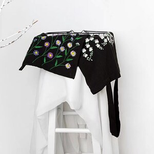 May include: Black apron with embroidered floral design. The apron features a black background with colorful embroidered flowers and green stems. The apron is hanging on a black metal rack over a white surface.