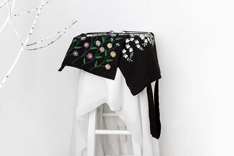 May include: Black apron with embroidered floral design. The apron features a black background with colorful embroidered flowers and green stems. The apron is hanging on a black metal rack over a white surface.