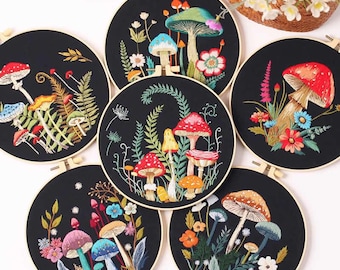 Pode incluir: Seis obras de arte de cogumelos bordados em tecido preto, cada uma em um bastidor de madeira. Os cogumelos são de várias cores, incluindo vermelho, azul e laranja, com costuras detalhadas de flores e folhagens. Os bastidores estão dispostos em uma superfície branca.