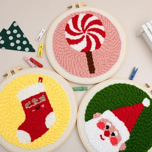 Op de afbeelding: Drie borduurringen met kerstthema. Eén toont een rood-witte swirl lolly op een roze achtergrond. Een andere toont een rode kous op een gele achtergrond. De derde toont de kerstman op een groene achtergrond.