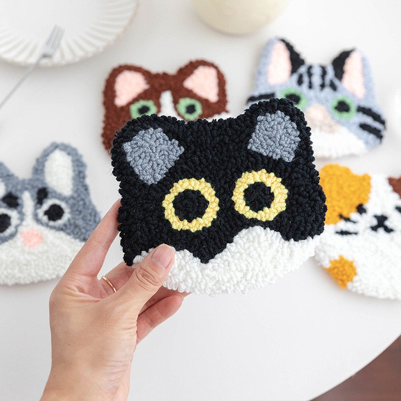 May include: Four colorful cat-shaped needle felted coasters. The coasters are made of wool and have different colored fur, including black, gray, brown, and orange. The coasters have black eyes with yellow centers.