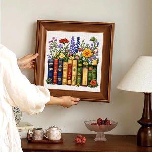 May include: A framed cross-stitch embroidery featuring a row of colourful books with vibrant wildflowers growing out of the top. The books are rendered in shades of blue, red, yellow, and green, while the flowers include purple, red, and yellow blooms.