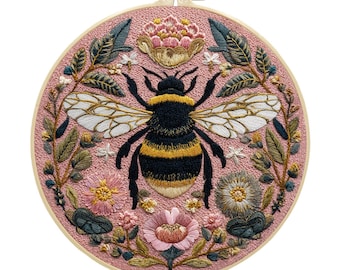 May include: A hand-stitched embroidery hoop featuring a detailed bumblebee design. The bee is black and yellow, with white wings, surrounded by floral and leaf embroidery in shades of pink, green, and cream. The hoop is a light pink color.