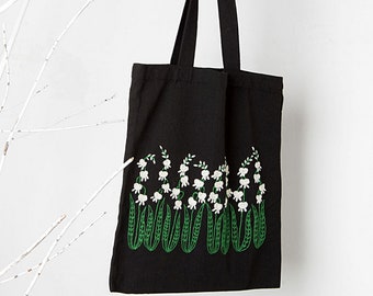May include: Black tote bag with a white and green embroidered design of lily of the valley flowers. The bag is hanging from a white branch and a black wire basket is in the foreground.