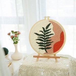 Pode incluir: Um aro de bordado de madeira com fundo branco e um desenho de samambaia verde. O aro est&aacute; em um suporte de madeira e tem um design abstrato rosa e laranja.