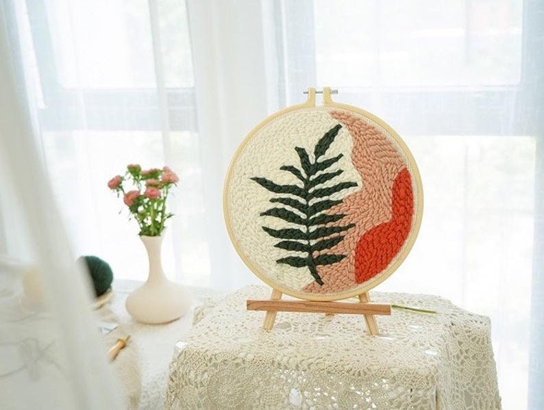 Pode incluir: Um aro de bordado de madeira com fundo branco e um desenho de samambaia verde. O aro est&aacute; em um suporte de madeira e tem um design abstrato rosa e laranja.