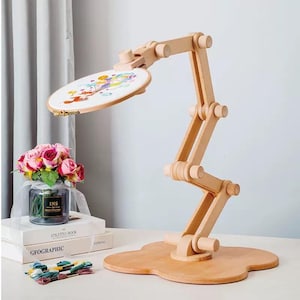 May include: A light-coloured wooden embroidery stand with an adjustable articulated arm and a circular base. A round embroidery hoop containing white fabric with a colourful printed design is attached to the top of the wooden stand.