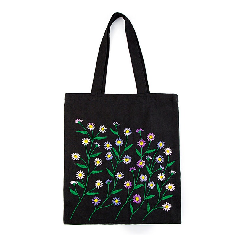 May include: Black tote bag with embroidered floral design. The flowers are white, yellow, and purple with green stems and leaves. The tote bag has a rectangular shape and two black handles.