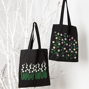 May include: Two black tote bags with floral embroidery. One bag features white lily of the valley flowers with green stems, while the other showcases a variety of colorful daisies. Both bags have long black handles.