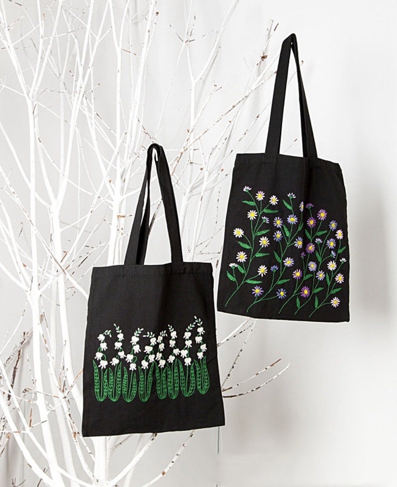 May include: Two black tote bags with floral embroidery. One bag features white lily of the valley flowers with green stems, while the other showcases a variety of colorful daisies. Both bags have long black handles.