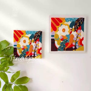 May include: Two square wall art pieces with colorful floral designs. The artwork features white flowers, orange, yellow, and blue accents, and is framed in white. The text "Tina House" is visible on the lower left of the image.
