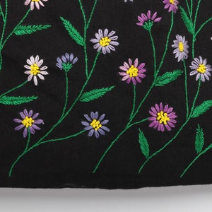 May include: Black fabric with embroidered floral design. The embroidery features daisies in shades of white, purple, and pink with yellow centers. Green stems and leaves complete the floral pattern.