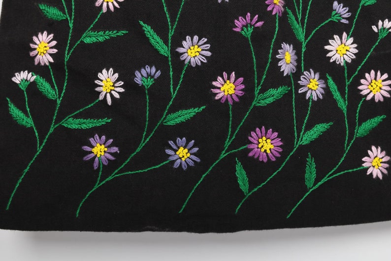 May include: Black fabric with embroidered floral design. The embroidery features daisies in shades of white, purple, and pink with yellow centers. Green stems and leaves complete the floral pattern.