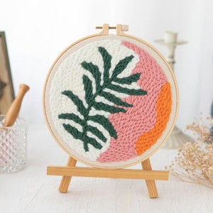 Pode incluir: Um aro de bordado redondo com uma moldura de madeira e uma tela branca. A tela apresenta um design de folha verde com detalhes rosa e laranja.