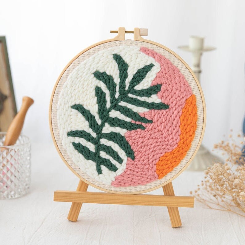 Pode incluir: Um aro de bordado redondo com uma moldura de madeira e uma tela branca. A tela apresenta um design de folha verde com detalhes rosa e laranja.