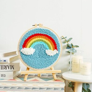 May include: A rainbow-coloured yarn art piece with white clouds, mounted on a wooden hoop and easel. The art piece is on a white background.