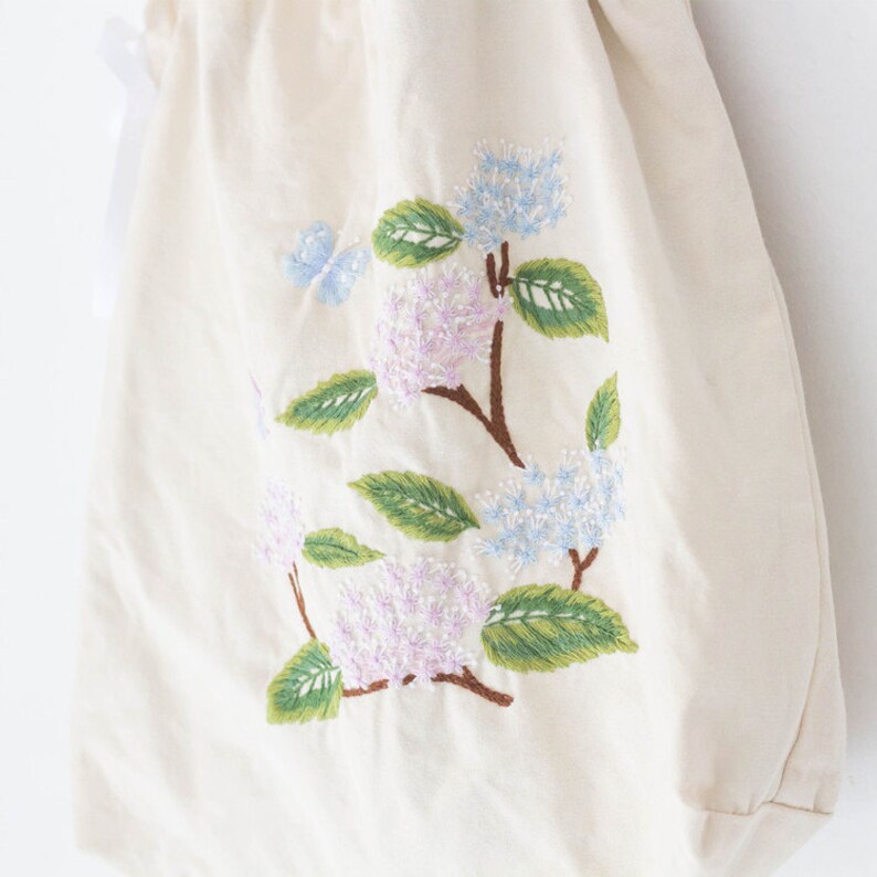 May include: A white canvas tote bag with an embroidered design of pink, blue, and green hydrangeas and a butterfly.