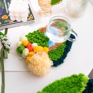 May include: A colorful, handmade coaster with a variety of textures and pom-poms. The coaster is green, blue, orange, and yellow. A glass of water is sitting on top of the coaster.