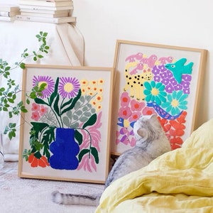 Könnte beinhalten: Zwei gerahmte Stickbilder mit einem farbenfrohen Blumen- und Tierdesign. Das Blumenstück zeigt eine blaue Vase mit rosa, orangefarbenen und grünen Blumen. Das Tierstück zeigt einen Leoparden, Vögel und Fische in einem Feld aus lila, blauen und gelben Blumen.