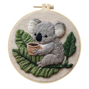 May include: Embroidered artwork showing a grey koala holding a small brown cup, sitting on a green leaf. The koala has a black nose and is surrounded by green leaves. The embroidery is set in a round wooden hoop.