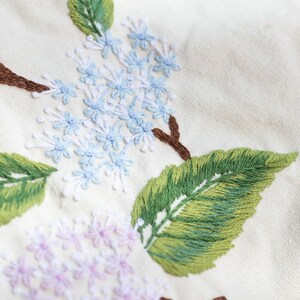 May include: Close-up of embroidered flowers on a white fabric. The flowers are light blue and pink, with green leaves. The embroidery is detailed and intricate.