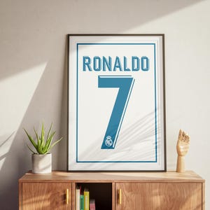 May include: Framed print displaying "RONALDO" and the number "7" in blue on a white background, with a team logo. The artwork is displayed on a wooden cabinet, alongside a potted plant and a wooden hand sculpture. The frame is black.
