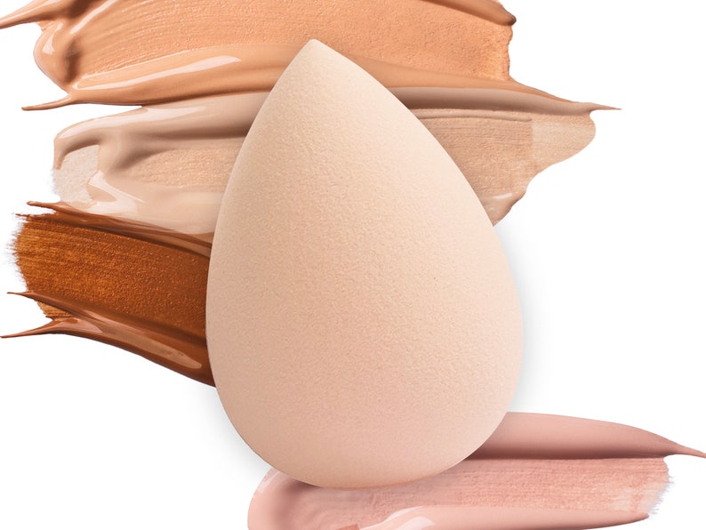 K&ouml;nnte beinhalten: Ein hellbeiger Make-up-Schwamm in Tropfenform, der auf drei Farbmuster von Foundation liegt. Die Farbmuster sind in den Farben hellbeige, mittelbeige und dunkelbraun.