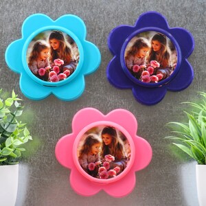 May include: Three flower-shaped photo frames against a white background. Each frame is a different colour: blue, purple, and pink. Each frame features a picture of two girls holding flowers.