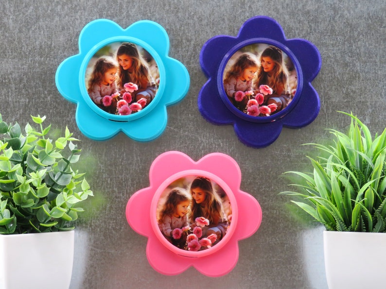 May include: Three flower-shaped photo frames against a white background. Each frame is a different colour: blue, purple, and pink. Each frame features a picture of two girls holding flowers.
