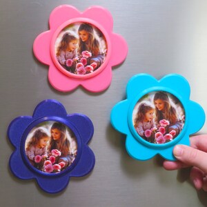 May include: Three flower-shaped photo frames with magnets on the back. Each frame is a different colour: pink, blue, and purple. Each frame has a photo of two girls holding flowers.