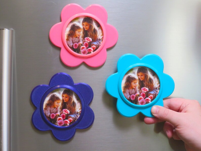 May include: Three flower-shaped photo frames with magnets on the back. Each frame is a different colour: pink, blue, and purple. Each frame has a photo of two girls holding flowers.