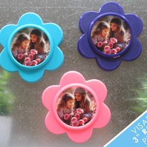 May include: Three flower-shaped photo frames in blue, pink, and purple, each with a 7.6 cm round photo insert. The photos show two young girls smiling and looking at flowers.