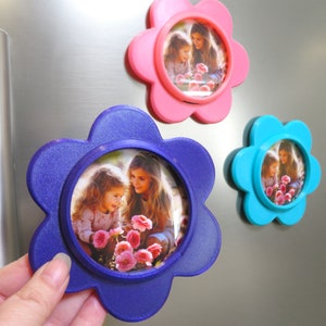 May include: Three flower-shaped photo frames with magnets on the back. The frames are pink, blue, and purple. Each frame has a photo of two girls looking at flowers.