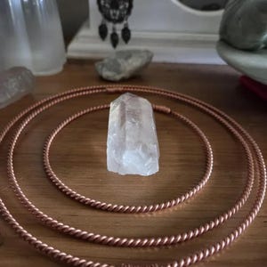 Könnte beinhalten: Eine klare, hexagonale Quarzkristallspitze, die in der Mitte von drei konzentrischen Kreisen aus gedrehtem Kupferdraht liegt. Der Kristall hat eine durchscheinende, facettierte Oberfläche und steht aufrecht auf einer glatten, hellbraunen Holzoberfläche.