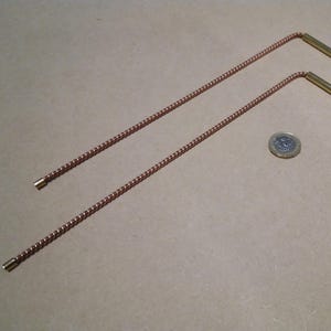 Puede incluir: Dos varillas metálicas anguladas con tallos trenzados de color cobre y extremos dorados. Una moneda se coloca entre ellas. Las varillas son probablemente herramientas o componentes.