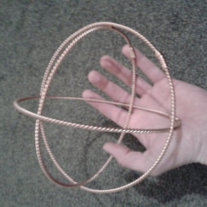 Könnte beinhalten: Eine goldfarbene Metalldrahtsphäre mit zwei sich schneidenden Kreisen. Die Kugel wird in der Hand einer Person gehalten.