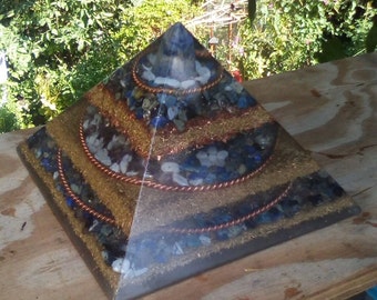 May include: A multi-layered crystal pyramid with copper wire accents. The pyramid is made of clear resin and contains various blue and white crystals. The pyramid is sitting on a wooden surface.