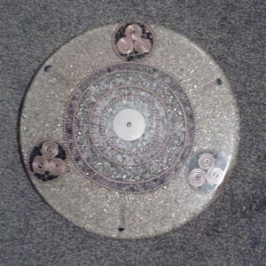 Op de afbeelding: Een ronde, witte, decoratieve plaat met een zilveren en witte kern en drie koperen draad triquetras rond de rand.