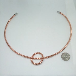 May include: A rose gold twisted wire choker necklace with a knotted circle pendant. The necklace is shown on a white surface with a British coin for scale.