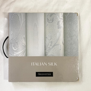 May include: A square sample book titled ITALIAN SILK by BREWSTER featuring four vertical panels of light gray wallpaper. The panels display varying textures including smooth surfaces, subtle wood grain patterns, and intricate embossed floral motifs.