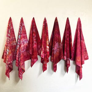 May include: Seven red and pink tie-dye fabric cloths hanging against a white wall. The cloths are draped, showcasing a variety of patterns and shades of red, pink, and hints of blue. The fabric appears soft and lightweight.