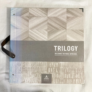 May include: A square sample book with a dark brown leather handle on the left side. The cover features a geometric wood grain pattern on top, a gray middle section with the text TRILOGY RECLAIMED. RESTORED. REFRESHED., and a textured bottom.