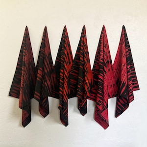 May include: Six red and black patterned fabric squares are displayed against a white wall. The fabric has a tie-dye or batik style design with vertical black stripes on a red background. The fabric is folded and hung, creating a decorative display.