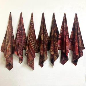 May include: Seven tie-dye fabric pieces in shades of red, brown, and tan are displayed against a white wall. Each piece is folded and hung, showcasing unique patterns and color variations. The textiles appear to be lightweight and suitable for various uses.