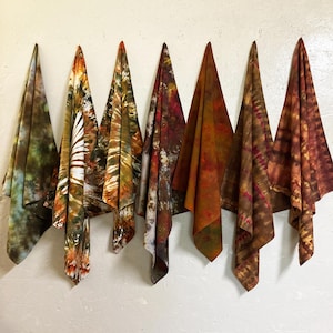 Puede incluir: Seis piezas de tela teñidas con la técnica tie-dye cuelgan contra una pared blanca. Las telas muestran una variedad de colores, incluyendo verdes, marrones, naranjas y rojos. Cada pieza tiene un patrón único, con diseños a rayas o marmolados.