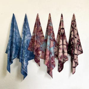 May include: Five tie-dye fabric pieces hanging against a white wall. The fabrics display a range of colors, including shades of blue, burgundy, and pink. The tie-dye patterns vary, creating a unique visual texture. The fabrics are likely towels or cloths.