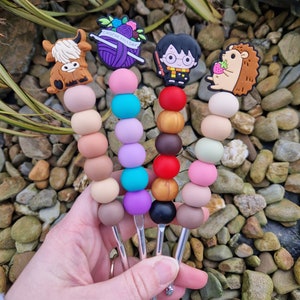 May include: Four colorful crochet hooks with different themed toppers. The toppers include a highland cow, a ball of yarn with flowers and the text "Croche Tally", Harry Potter, and a hedgehog with a strawberry.
