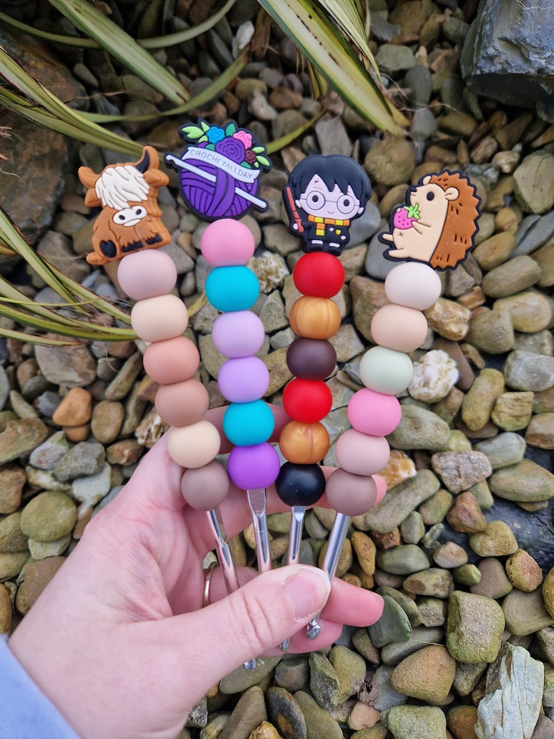 May include: Four colorful crochet hooks with different themed toppers. The toppers include a highland cow, a ball of yarn with flowers and the text "Croche Tally", Harry Potter, and a hedgehog with a strawberry.