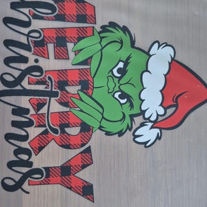 May include: A green Grinch character wearing a red Santa hat with the words "Merry Christmas" in black and red buffalo plaid.